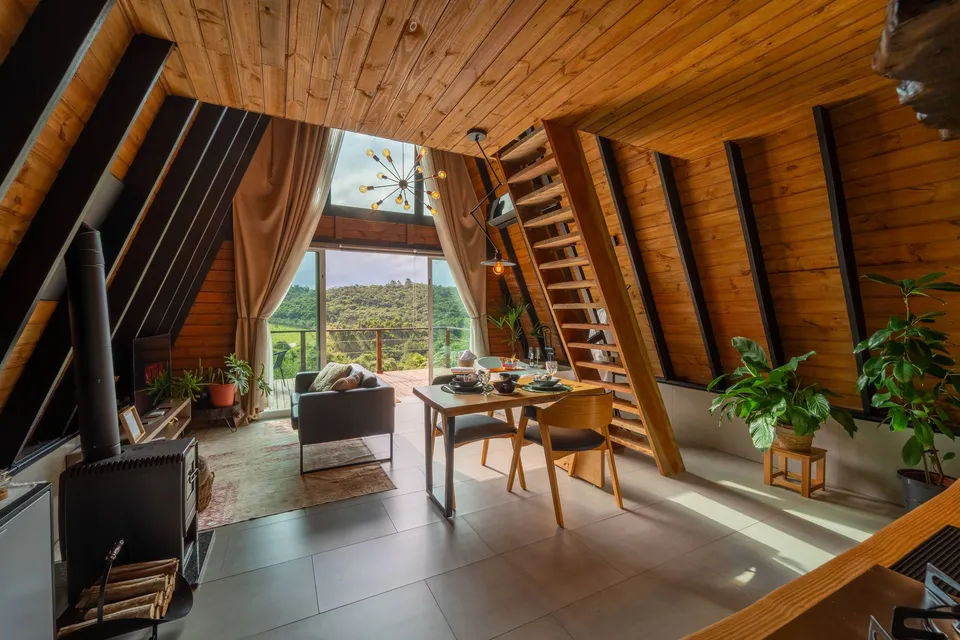 Sala do chalé com lareira, mesa posta e a janela alta aberta para o verde